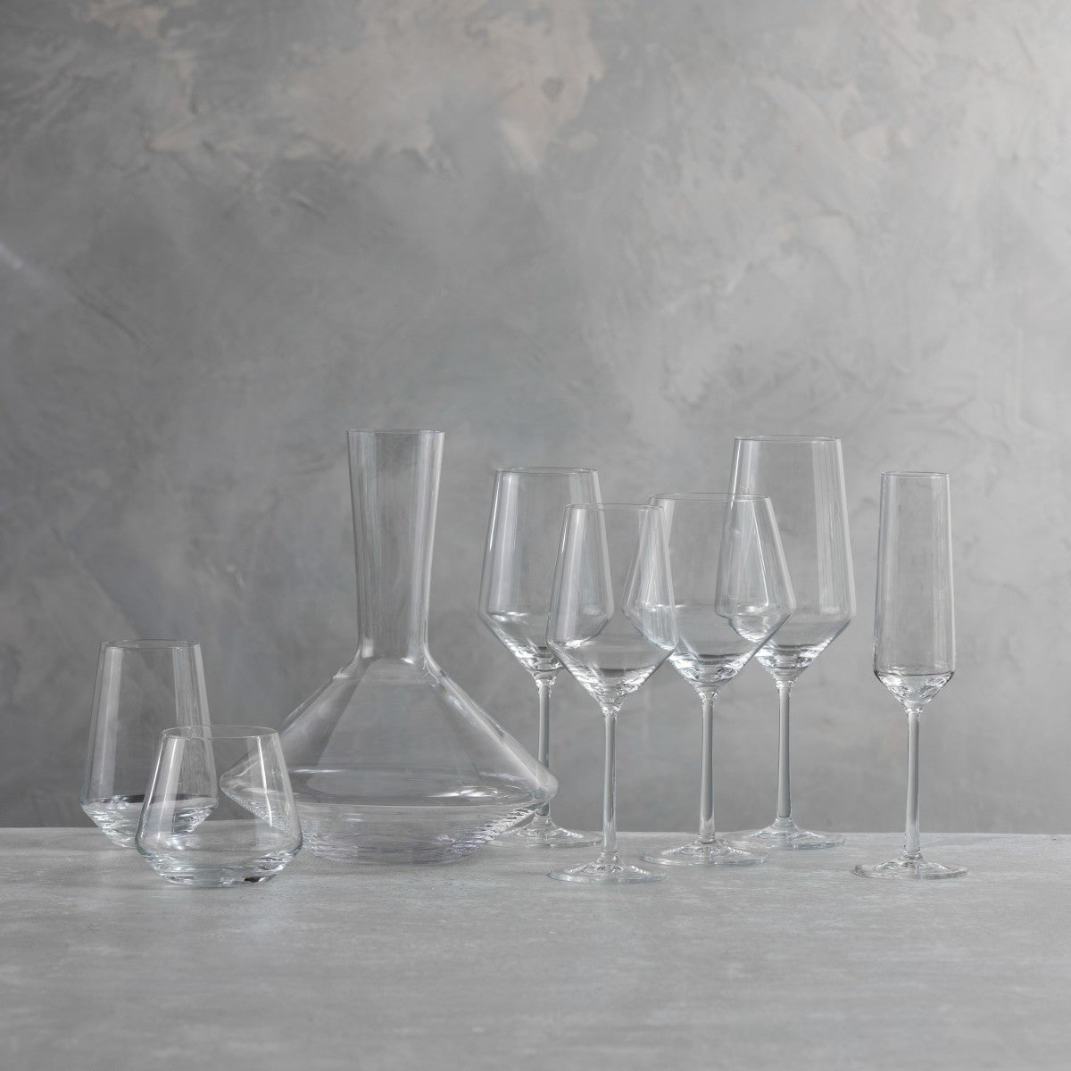 The Zwiesel Fortessa Zweisel Pure Barware set, made of durable Tritan, features a decanter, two stemless wine glasses, three wine glasses, and two champagne flutes arranged on a light surface against a textured gray background.