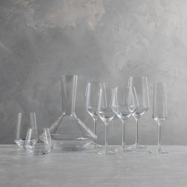 The Zwiesel Fortessa Zweisel Pure Barware set, made of durable Tritan, features a decanter, two stemless wine glasses, three wine glasses, and two champagne flutes arranged on a light surface against a textured gray background.