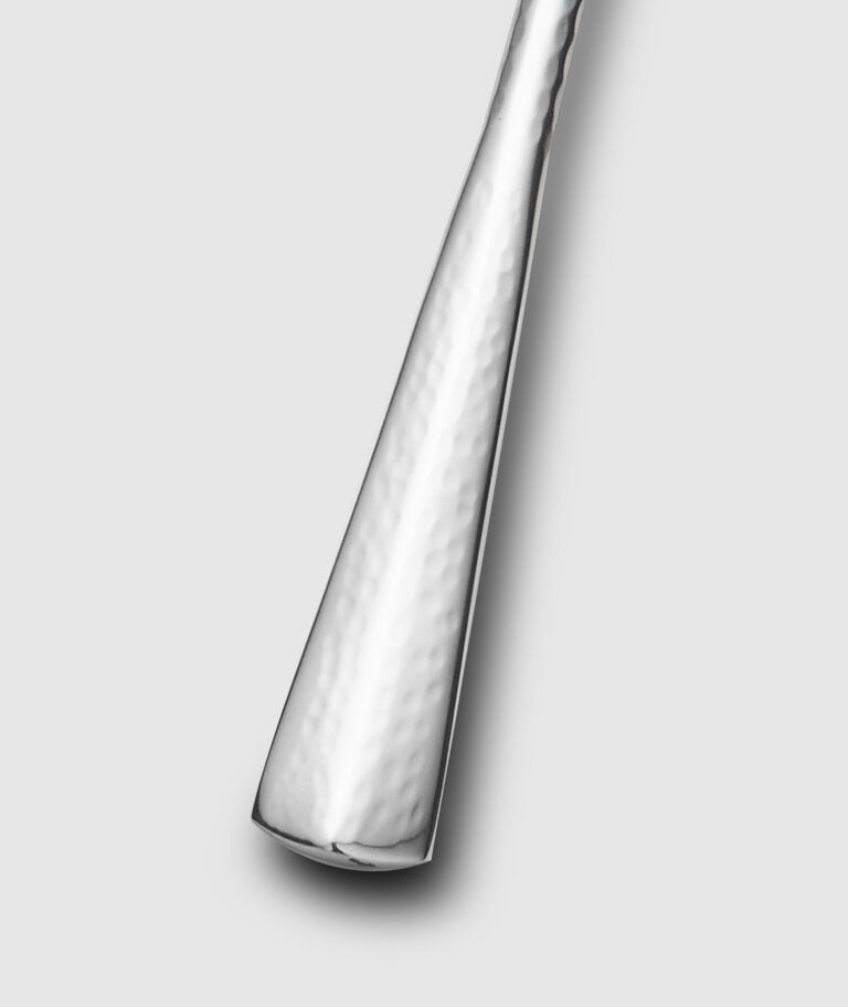A close-up of the Mary Jurek Alta Slotted Serving Spoon handle highlights its hammered, textured finish against a light gray background—an elegant upgrade to your stainless steel serveware.