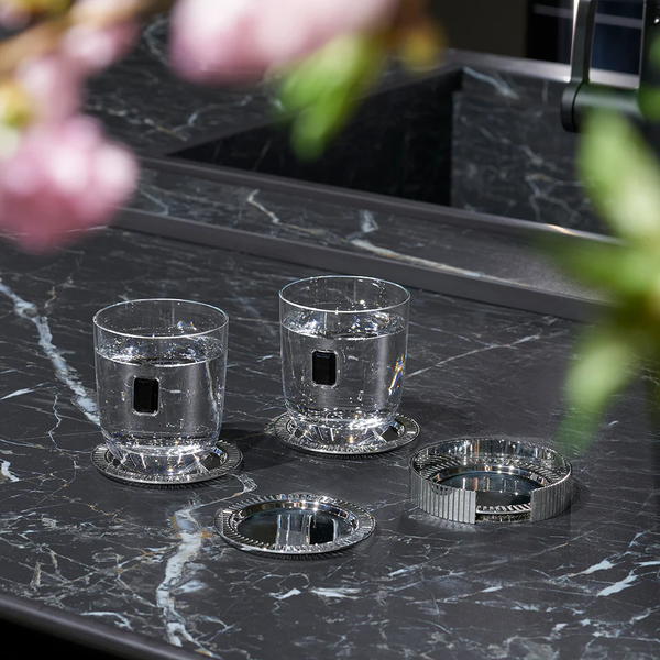 Two clear glasses of water with square black accents sit on a dark marble countertop, each resting on a Striations Silver Coaster from Anna New York. Blurred pink flowers and green leaves appear in the foreground.