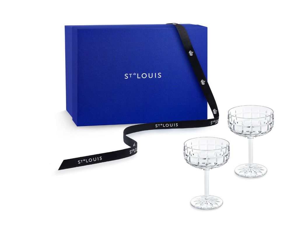 A royal blue Saint-Louis Manhattan Barware gift box with white text and a black ribbon is shown, featuring two clear, patterned crystal cocktail glasses on stems in front.