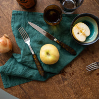 A Sabre Tortoise Flatware 5 Piece Setting sits on a green napkin with a yellow apple, halved apple in a bowl, onion, and an empty fork arranged on a wooden table.