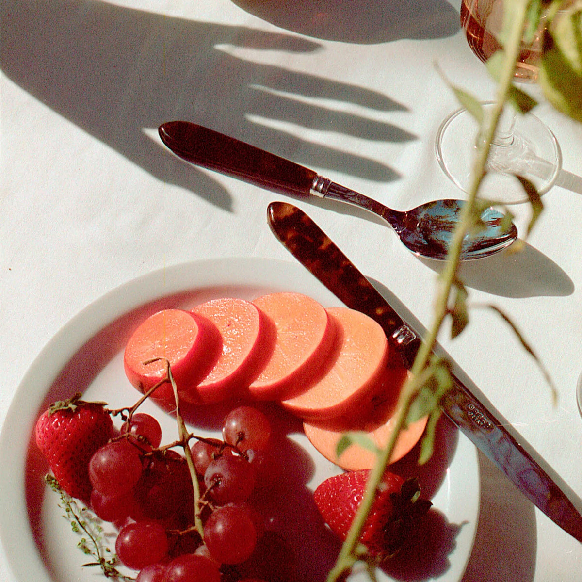 A plate with pink sausage, strawberries, and red grapes sits on a white tablecloth. Nearby, Sabre Tortoise Flatware 5 Piece Setting with 18/10 stainless steel blade and acrylic handle gleams beside a shadowed hand and green plant stems.