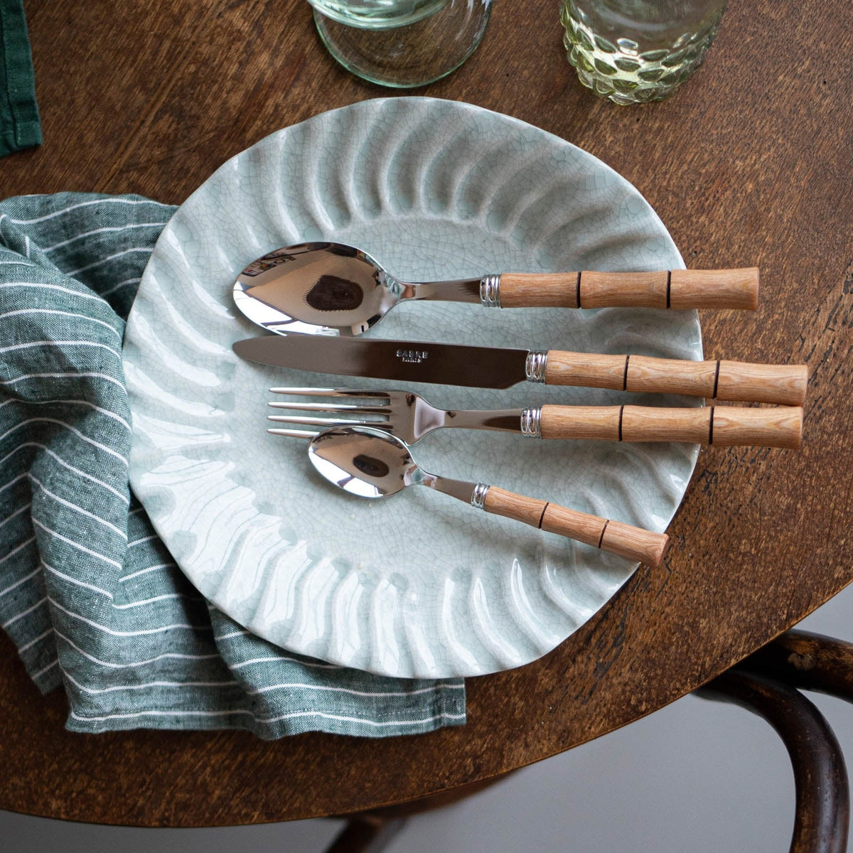 A ceramic plate with a wavy pattern sits on a wooden table, holding the Sabre Bamboo Flatware 5 Piece Setting. A striped green napkin peeks out from under the plate, with glasses nearby.