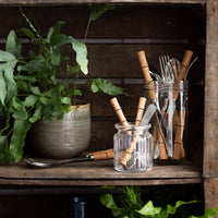 A rustic wooden shelf holds a potted fern and glass jars with Sabre Bamboo Flatware 5 Piece Setting, featuring bamboo-style handles, surrounded by green foliage.