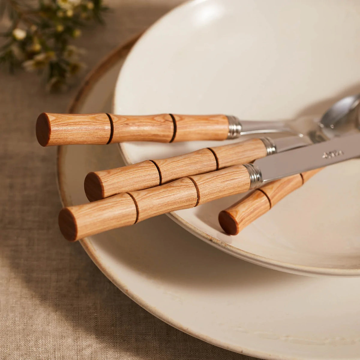 A Sabre Bamboo Flatware 5 Piece Setting rests on stacked white ceramic plates atop a beige tablecloth, with blurred greenery in the background.