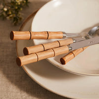 A Sabre Bamboo Flatware 5 Piece Setting rests on stacked white ceramic plates atop a beige tablecloth, with blurred greenery in the background.