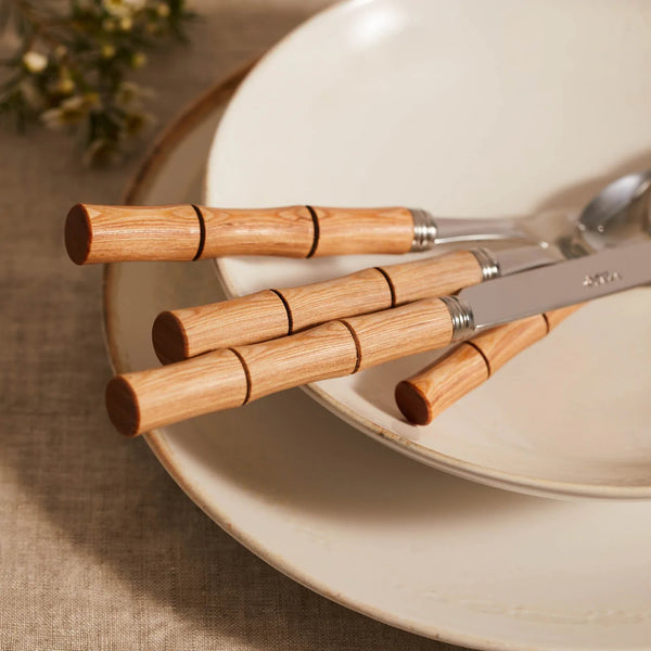A Sabre Bamboo Flatware 5 Piece Setting rests on stacked white ceramic plates atop a beige tablecloth, with blurred greenery in the background.