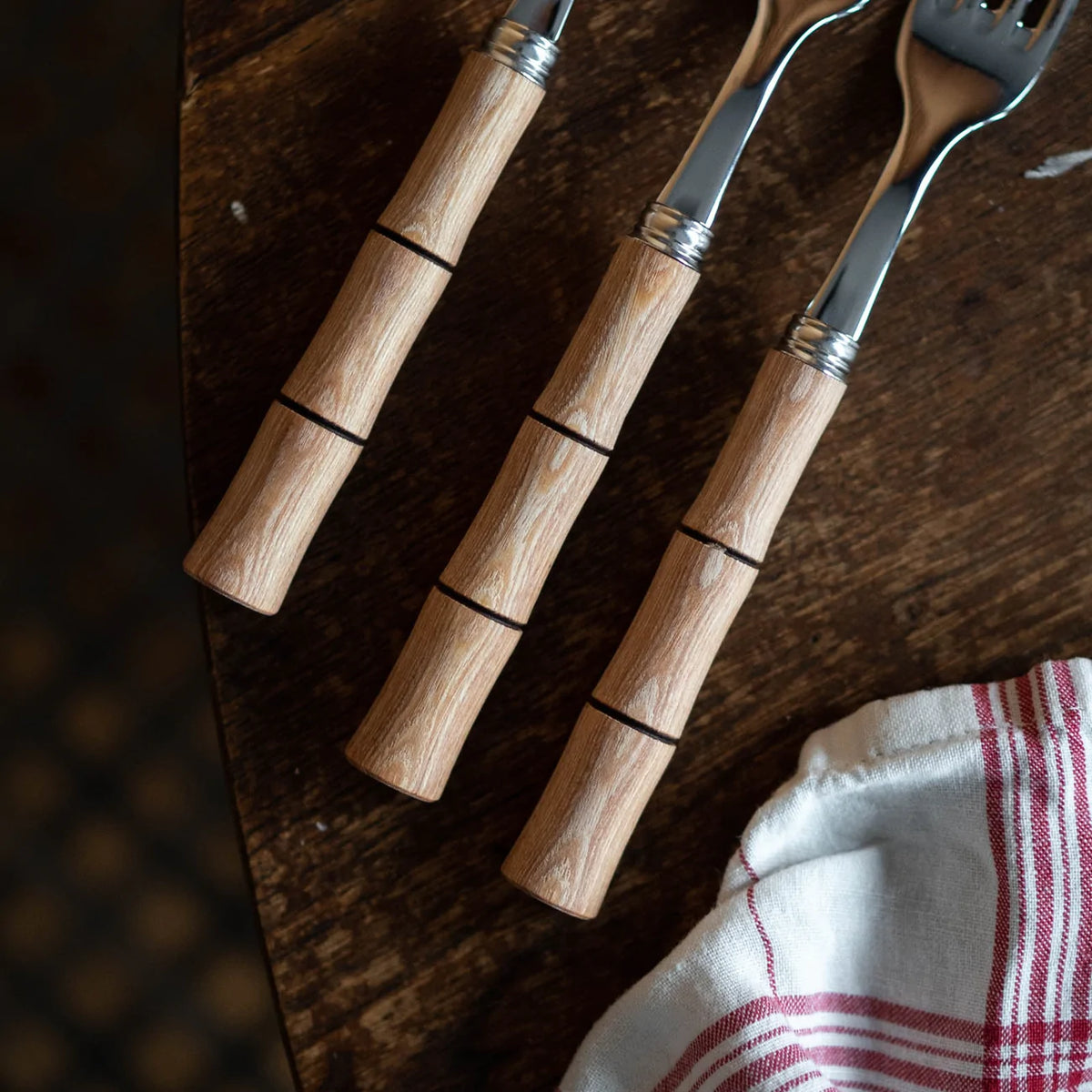 Three silver forks from the Sabre Bamboo Flatware 5 Piece Setting, featuring light wooden bamboo-patterned handles, rest on a dark wood table beside a white cloth with red stripes. The photo is taken from above.