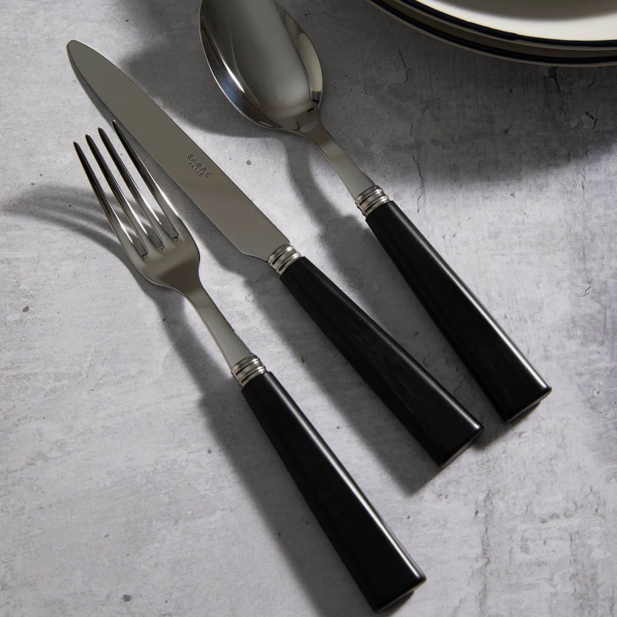 The Sabre Nature Flatware 5 Piece Setting in Black Wood features a fork, knife, and spoon with shiny stainless steel heads and black handles, neatly arranged on a light gray textured surface beside stacked plates.