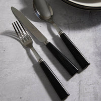 The Sabre Nature Flatware 5 Piece Setting in Black Wood features a fork, knife, and spoon with shiny stainless steel heads and black handles, neatly arranged on a light gray textured surface beside stacked plates.