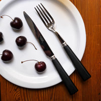 A white plate with four cherries, a fork, and a knife with black handles from the Sabre Nature Flatware 5 Piece Setting - Black Wood, on a wooden table.