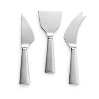 The Simon Pearce Hartland Cheese Knife Set features three silver stainless steel knives with wide handles—pointed, flat and wide, and curved with serrated edge and forked tip—elegantly arranged on a white background.