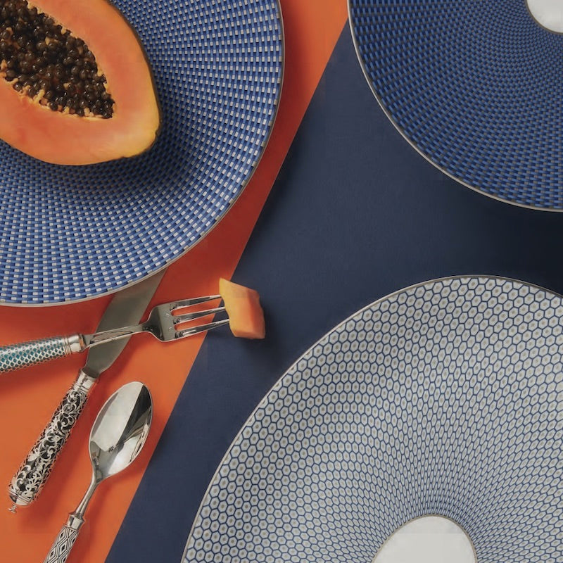 A papaya half sits on a Raynaud Tresor Dinnerware Blue plate, beside a fork with papaya, knife, and spoon, all atop orange and navy blue tablecloths accented by more formal Raynaud Tresor dinnerware.