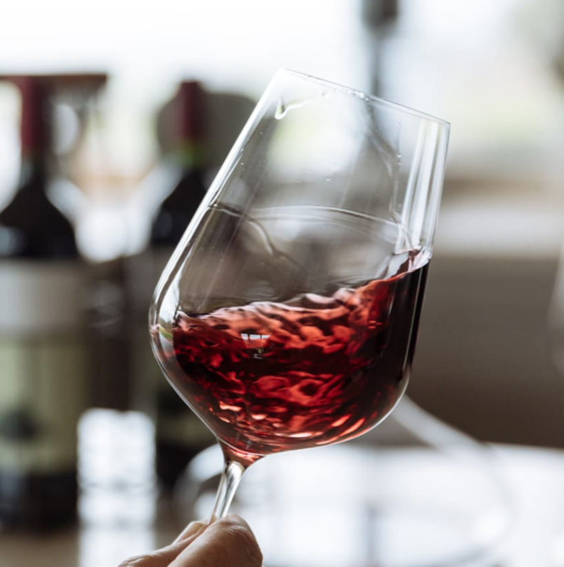 A close-up of a hand swirling red wine in a Richard Brendon Jancis Robinson Universal Wine Glass, with blurred bottles and a bright window behind; part of the Set of 2.