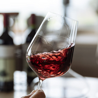 A close-up of a hand swirling red wine in a Richard Brendon Jancis Robinson Universal Wine Glass, with blurred bottles and a bright window behind; part of the Set of 2.