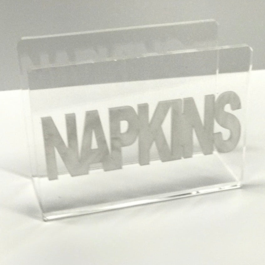 The Eye Candy Acrylic Napkin Holder features bold silver "NAPKINS" lettering across the front, set against a plain, light-colored background.