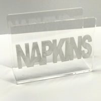 The Eye Candy Acrylic Napkin Holder features bold silver "NAPKINS" lettering across the front, set against a plain, light-colored background.
