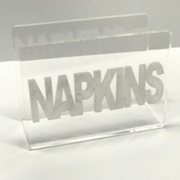 The Eye Candy Acrylic Napkin Holder features bold silver "NAPKINS" lettering across the front, set against a plain, light-colored background.