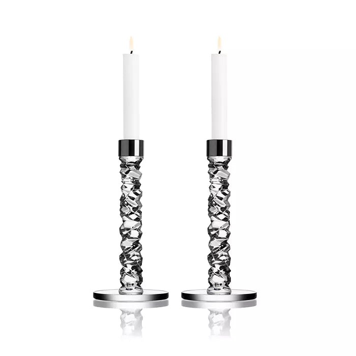 Two tall white candles glow in Orrefors Kosta Boda Carat Candlesticks Silver Set of 2, with distinctive twisted designs and round bases. The elegant crystal craftsmanship is highlighted against a plain white background.