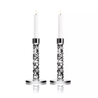 Two tall white candles glow in Orrefors Kosta Boda Carat Candlesticks Silver Set of 2, with distinctive twisted designs and round bases. The elegant crystal craftsmanship is highlighted against a plain white background.