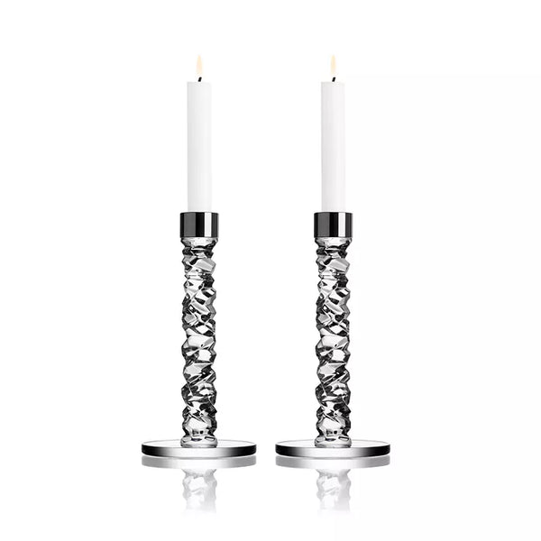 Two tall white candles glow in Orrefors Kosta Boda Carat Candlesticks Silver Set of 2, with distinctive twisted designs and round bases. The elegant crystal craftsmanship is highlighted against a plain white background.