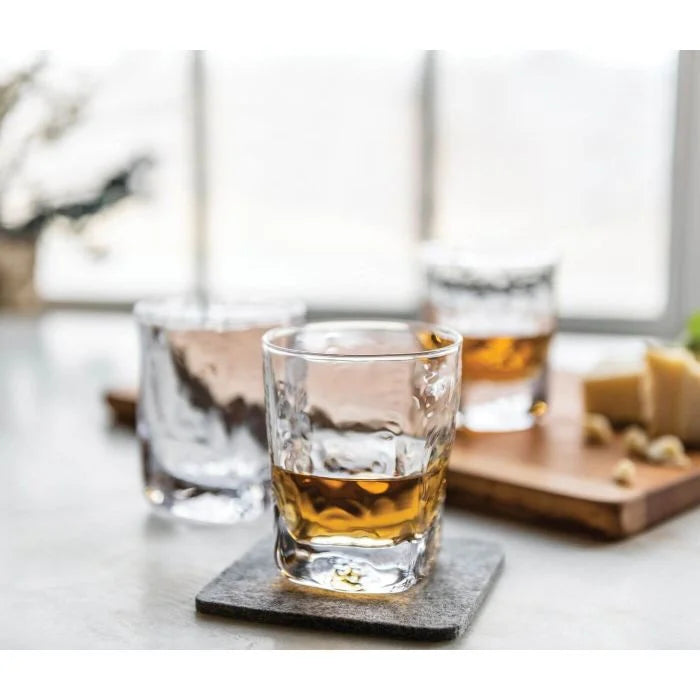 A clear Simon Pearce Woodbury Bourbon glass, filled with amber liquid, rests on a coaster with two more glasses and a wooden cheese-and-nut board softly blurred in the background on a light surface.
