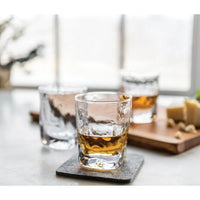 A clear Simon Pearce Woodbury Bourbon glass, filled with amber liquid, rests on a coaster with two more glasses and a wooden cheese-and-nut board softly blurred in the background on a light surface.