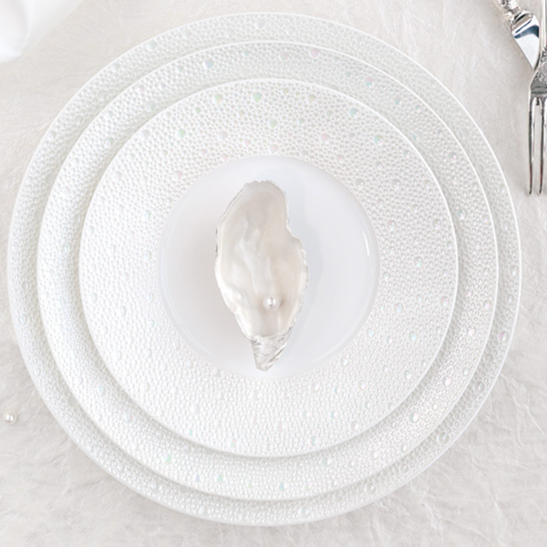 A white oyster shell with a pearl sits on a Bernardaud Ecume Dinnerware Perle plate, layered over two larger textured plates, with a fork and knife at the top right, all set against a pristine white background.