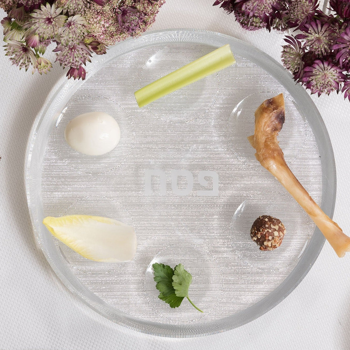 The Apeloig Acrylic Seder Plate showcases six symbolic foods—boiled quail egg, celery stick, chicken drumette bone, brown truffle, parsley sprig, and endive piece—on a white tablecloth with nearby purple flowers.
