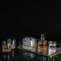 A gold tray holds a glass decanter with brown liquor, a glass ice bucket, and a small pitcher. Two Richard Brendon Diamond DOF glasses, one filled, sit beside the tray on dark marble against a black background.