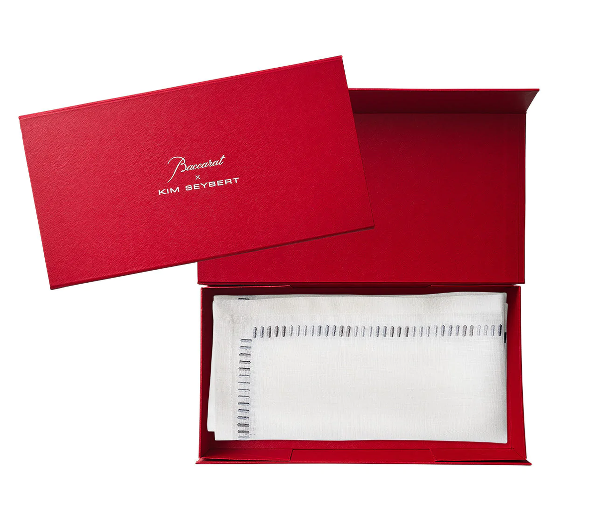 A red rectangular box with "Baccarat x Kim Seybert" on the lid opens to reveal a neatly folded Harmonie Napkin in White, Gray & Silver, Set of 4 by Kim Seybert, each featuring a stitched border.