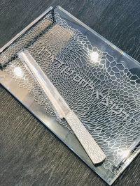 The Organic Lace Challah Knife Silver by Metalace Art, with a textured handle, rests on a rectangular clear tray featuring an intricate geometric metal lace art pattern and partially visible engraved text on a dark surface.