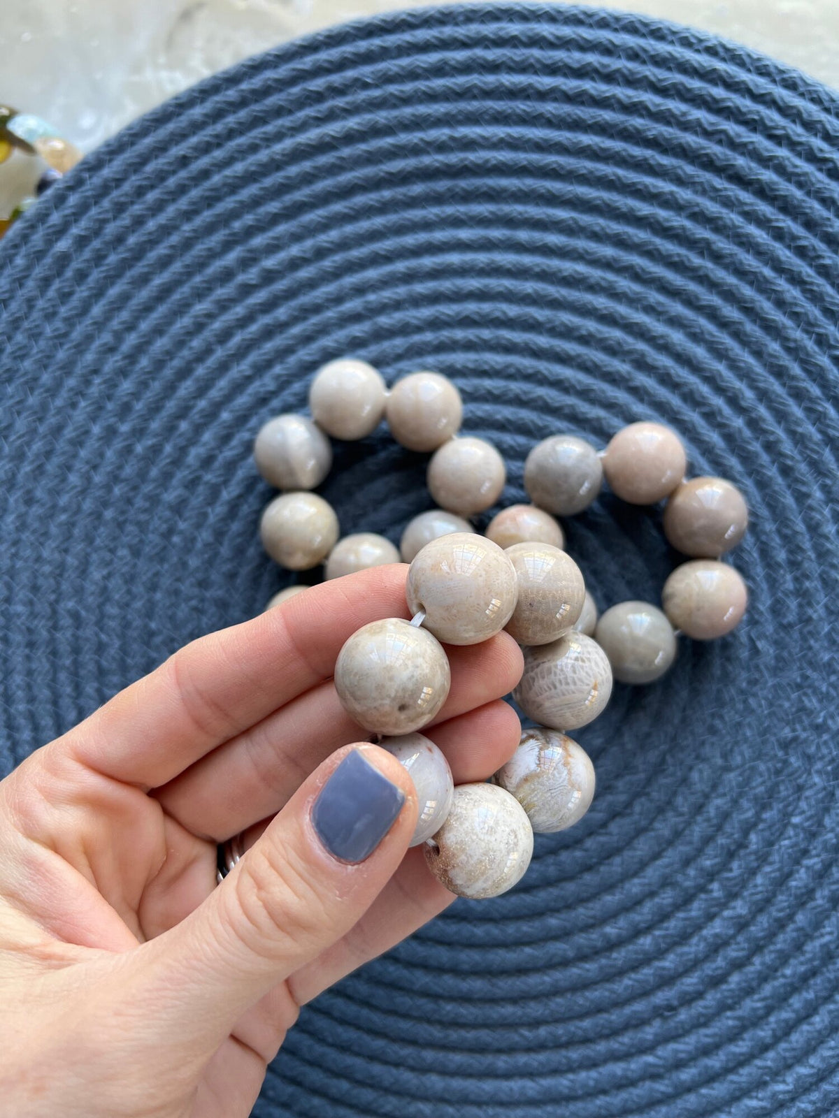 A hand with painted nails holds a Whigstock Studio Marble Bead Napkin Ring Cream (Set of 4) over a textured blue mat, with the rest of the natural stone napkin rings arranged in a heart shape to showcase their handmade beauty.