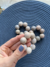 A hand with painted nails holds a Whigstock Studio Marble Bead Napkin Ring Cream (Set of 4) over a textured blue mat, with the rest of the natural stone napkin rings arranged in a heart shape to showcase their handmade beauty.