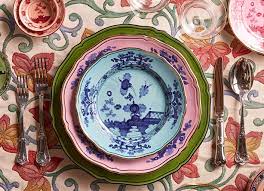 A vibrant table setting features Ginori 1735's Oriente Italiano Dinnerware Iris, with plates stacked in blue, pink, and green chinoiserie florals, surrounded by cutlery and glasses on a floral tablecloth.