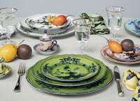 A table set with Ginori 1735’s Oriente Italiano Dinnerware Malachite, glasses of water, silverware, napkins, and assorted fruits like pears and plums creates an elegant and eclectic Italian tableware display.