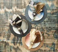 Three plates with patterned napkins and spiked napkin rings are arranged on Kim Seybert’s Croco Placemat Charcoal Set of 4, displayed atop a blue and brown tie-dye tablecloth. The plates and napkins coordinate in blue, brown, and gray tones.