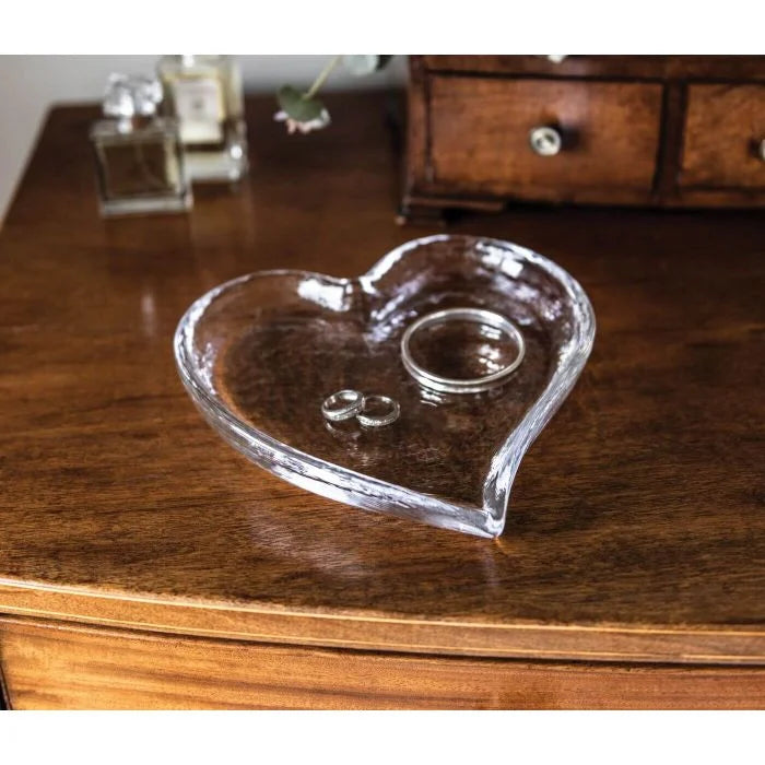 A Simon Pearce Heart Tray holds three silver rings on a wooden dresser, with perfume bottles and a jewelry box in the background—an elegant gift idea.