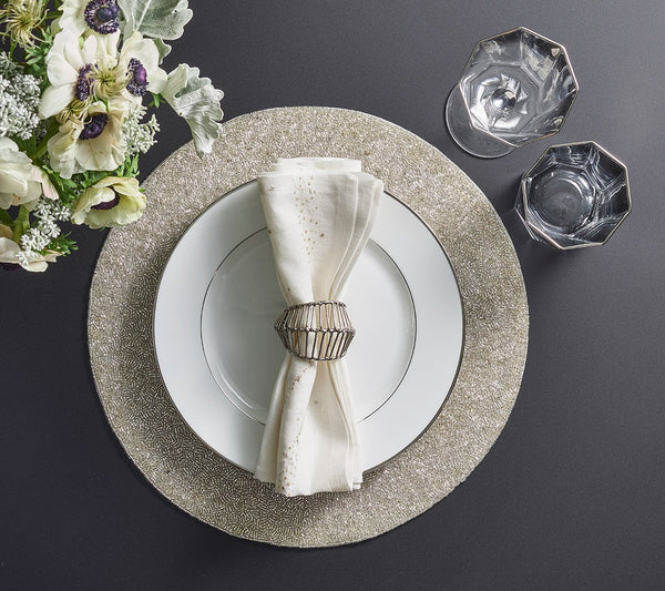 Create an elegant table setting with the Kim Seybert Star Spray Napkin White & Gold & Silver Set of 4, paired with a white plate on a silver charger, gold napkin ring, faceted glasses, and a floral arrangement on a dark surface.