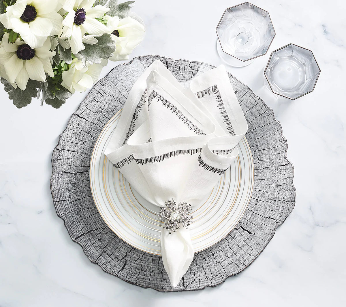 An elegant table setting featuring Kim Seybert's Filament Napkin White & Gunmetal Set of 4, paired with a silver-rimmed plate, textured charger, jeweled napkin ring, two clear glasses, and white flowers on a marble surface.