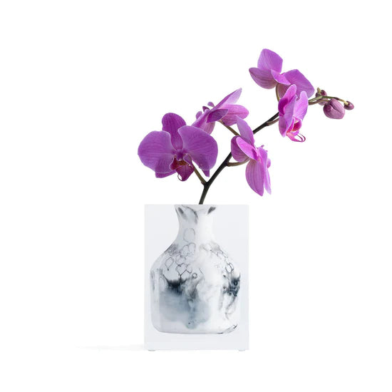 A vase from the JR William Lucite Vase Collection with a marbled black finish holds vibrant purple orchids against a plain white background, adding a touch of modern luxury.