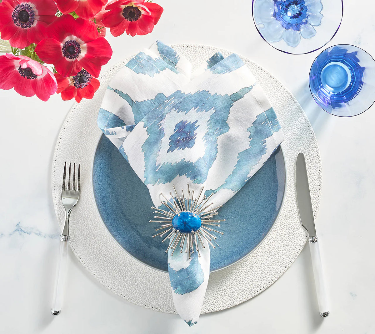 A table is set with a blue plate on a white charger, fork and knife, blue glasses, and an indigo Kim Seybert Watercolor Ikat Napkin with a spiky blue napkin ring, plus a red flower bouquet in the corner.
