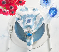 A table is set with a blue plate on a white charger, fork and knife, blue glasses, and an indigo Kim Seybert Watercolor Ikat Napkin with a spiky blue napkin ring, plus a red flower bouquet in the corner.