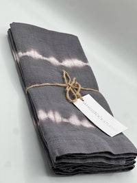 A folded, dark gray Hand Dyed Linen Napkin from Whigstock Studio's Set of 4 - Midnight, featuring white tie-dye stripes and twine, is displayed with a branded tag on a plain light background as a sustainable essential.