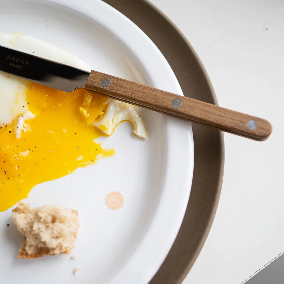 A fried egg with a broken yolk and a piece of bread sit on a white plate, accompanied by the Sabre Bistro Flatware 5 Piece Setting - Teak.
