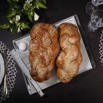 Two braided challah loaves with sesame seeds sit on the Apeloig Collection Metallic Silver Acrylic Challah Board, accompanied by a knife and salt dish, on a dark table with flowers, plates, cutlery, and glasses.