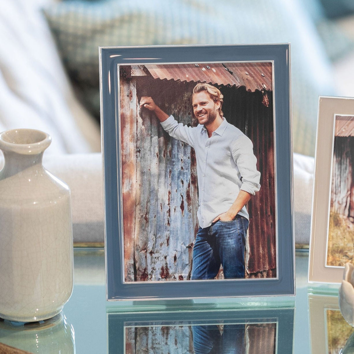 A framed photo in the Addison Ross Enamel Thin Frame - Denim, featuring a smiling man in a light blue shirt and jeans against a metal wall, is displayed on glass beside a white ceramic vase. The frame has elegant silver trim for a refined touch.