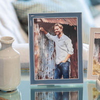 A framed photo in the Addison Ross Enamel Thin Frame - Denim, featuring a smiling man in a light blue shirt and jeans against a metal wall, is displayed on glass beside a white ceramic vase. The frame has elegant silver trim for a refined touch.