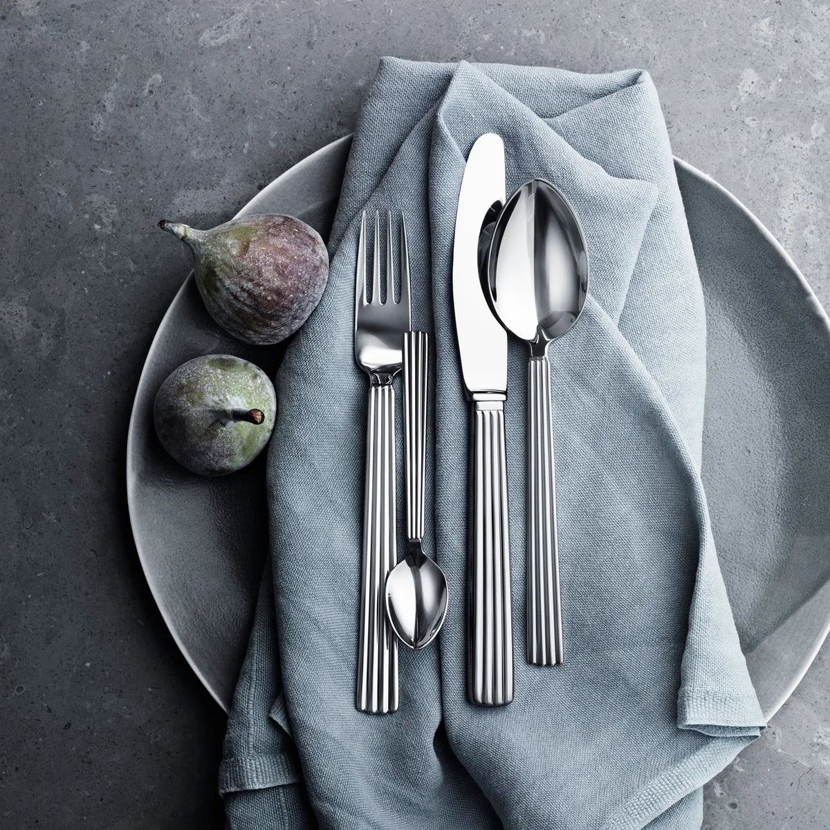 A plate with a folded gray cloth holds the Georg Jensen Bernadotte Flatware 5 Piece Setting—fork, knife, spoon, and teaspoon—with two figs beside the cutlery on a stone-like gray surface.
